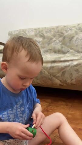 A young child sits barefoot on a tiled floor, threading colorful wooden blocks onto a red string. The child, wearing a blue t-shirt, focuses intently on the task, maneuvering the string through the holes in the blocks. Several blocks of various colors, including green, orange, pink, and yellow, are scattered around the child on the floor. A sofa with a floral pattern is visible in the background. The camera angle is relatively close, following the child's hands and the blocks.
