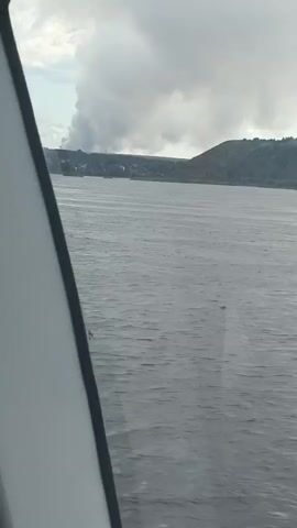 The video shows a wide shot of a body of water with a shoreline and hills in the distance. A large plume of smoke or steam rises from the land on the far side, obscuring the horizon and the sky. The water is rippled, indicating some movement on the surface. The foreground is partially obscured by a white object with a black edge, which appears to be part of a vehicle or structure. The overall impression is a hazy, somewhat dramatic landscape.