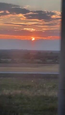 The video shows a sunset over a rural landscape. The sun is setting behind a band of clouds on the horizon, casting a warm glow across the sky. Below the sky, there is a wide expanse of fields and scattered trees. In the foreground, there is tall grass and some bushes. The image appears to be taken from a slightly elevated perspective, possibly from a vehicle or a hill. There are no people or visible objects in the immediate foreground. The overall scene is serene and natural.