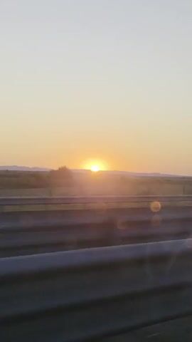 The video shows a sunset viewed from a moving vehicle. The sun is low on the horizon, casting a warm glow across the sky. In the foreground, there are two metal guardrails, indicating the vehicle is on a road, likely a highway. The landscape beyond the guardrails appears to be rural, with rolling hills and sparse vegetation. The sky is a gradient of pale yellow to soft orange, transitioning to a clear pale blue at the top of the frame. There are some lens flares visible, suggesting the sun's light is hitting the camera lens directly. In one brief segment, a large, empty billboard structure is visible against the sunset. Later, a car passes by in the opposite direction, its silhouette blurred due to its speed and the movement of the camera. The overall impression is one of a scenic journey during dusk.