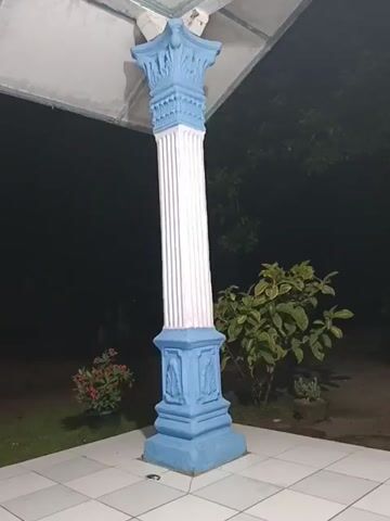 The video shows a decorative pillar, painted white and blue, standing on a tiled surface. In the background, there are plants and trees, and in the far distance, a dimly lit building can be seen. The lighting suggests it is nighttime. The camera appears to be positioned looking out from under a structure supported by the pillar. The video is static, with no movement or audio.