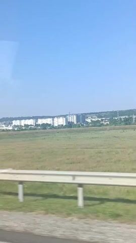 This is a video of a car or bus moving along a road, with a view of a rural landscape and a distant city. The road in the foreground has a white guardrail. Beyond the road is a large, grassy field. In the distance, there is a city with many tall, white buildings, possibly apartment blocks. The sky is a clear blue, suggesting it is daytime and the weather is good. The scene appears to be in a rural area outside of a city in Mitoc, Moldova. There is no visible text in the video. The overall mood is peaceful and unremarkable, likely capturing a journey.