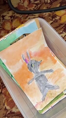The video shows a person's hands rummaging through a clear plastic container filled with children's artwork. The artworks are varied in style and subject matter, including a drawing of a gray rabbit with pink ears and large green eyes against an orange background, a drawing of a smiling person in a blue uniform with yellow hair and skin, and a drawing of a pink dress with colorful polka dots. The container is placed on a patterned surface, possibly a rug or carpet. The lighting is dim, and the focus shifts between the different drawings as the hands move them around. The video ends with a close-up of a drawing of a pink dress.