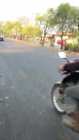 The media file shows a person riding a motorcycle on a paved road in Kludan, East Java. The rider, wearing a black helmet, a dark hoodie, and jeans, is positioned in the center of the frame, moving away from the camera. The motorcycle is red and silver. The scene is outdoors with trees lining the right side of the road and buildings visible in the background. Another motorcycle and a red car are also present on the road, suggesting traffic. The lighting indicates it is daytime, possibly late afternoon or early evening, with the sun casting a warm glow. The overall atmosphere appears calm, with no strong emotional cues evident. The visible text on the motorcycle's license plate is "S 6337 KB".