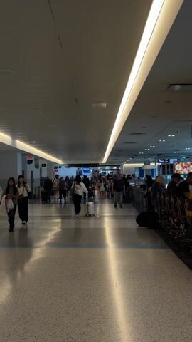 The video shows the interior of an airport terminal. Numerous people are walking in various directions, many pulling luggage. Some are standing near a seating area on the right side of the frame. The floor is light-colored tile, and the ceiling is bright with linear lighting fixtures. Signage indicates directions to different gates. The overall environment appears busy.