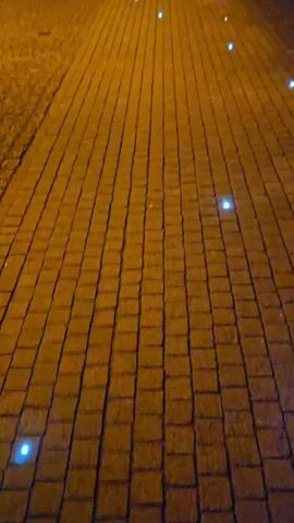 The video shows a brick-paved pathway at night illuminated by rows of small, blue lights embedded in the brickwork. The lights guide the path toward a building in the background. To the right of the pathway is a large, white wheel-shaped structure with mine carts positioned underneath. The structure is also illuminated. There's a tree partially obscuring the view of the structure. The overall ambiance is dark, highlighting the illuminated path and objects.
