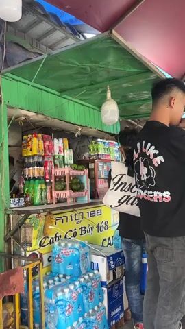 The video shows a group of young people gathered at a small, outdoor cafe stand. The stand is stocked with beverages and snacks, and has a green and yellow sign in front that reads "Cafe Cô Bay 168 CAFE". There are several people standing and sitting around a table under a large umbrella. One person is wearing a black t-shirt with a graphic design and grey jeans. Another is wearing a green hooded sweatshirt and wide-legged blue jeans. The cafe appears to be in a bustling area, with a covered walkway behind it. People are seated at the table and appear to be eating and drinking.