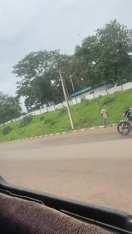 This is a video shot from the interior of a moving vehicle, likely a bus, looking out the window. The scene depicts a roadside in Aso, Nigeria. The time of day appears to be overcast and possibly raining, as the sky is a pale grey and the road surface is wet.

The foreground shows the edge of the vehicle's interior, with a brown, textured seat or trim. The middle ground is dominated by a paved road with a white and black striped curb. Beyond the curb, a grassy embankment slopes upwards. Various trees, some with dense green foliage, are visible on top of the embankment.

Several objects and people are present. A utility pole with wires stands prominently on the embankment. A motorcycle with two people riding it is seen on the road, heading in the same direction as the camera vehicle. Further along the road, a large billboard is visible, advertising "SALIS VENTILATED HOMES." The billboard includes a phone number, "09091122331," and a website address, "www.salishomes.org." Two people are walking on the sidewalk near the billboard. Another person is walking further down the road, on the sidewalk. A white vehicle, possibly a truck, is parked on the side of the road in the distance.

The overall impression is a typical urban or suburban street scene in Nigeria, possibly on a cloudy or wet day. The presence of the billboard suggests a commercial area. The activity is simply the passage of vehicles and pedestrians along the road. There are no strong emotional cues conveyed by the imagery.
