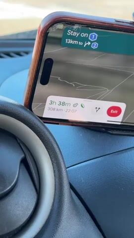 The video begins with a close-up of a smartphone displaying a navigation app on a car dashboard. The app shows a route with a remaining distance of 13 km and an estimated travel time of 3 hours and 38 minutes for a total distance of 308 km. The car appears to be in motion, as indicated by the blurred background and the presence of an air vent in the foreground. A hula dancer figurine is visible on the dashboard. The scene then shifts to a view of the rear of a red Opel car ahead, displaying a license plate that reads "WEN RE 200." The Opel is in traffic, possibly approaching a toll booth, as indicated by the overhead signs with traffic signals. The video captures a brief moment of a road trip, showcasing the in-car environment and the car ahead.