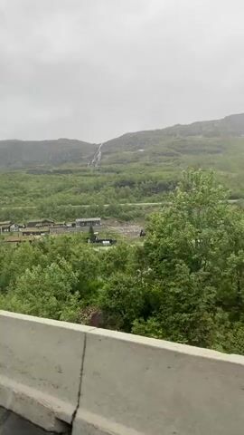 This media file captures a scenic view of a village nestled against a lush green mountainside in Røldal, Norway. The scene is dominated by natural elements: a towering mountain range, verdant slopes covered in trees and vegetation, and a thin waterfall cascading down the rocky face of the mountain in the distance.

The village itself consists of several small, dark-colored houses with traditional turf roofs, indicative of Norwegian architecture. These houses are spread out along the base of the mountain, some appearing to be clustered together. A road curves through the landscape, with a concrete barrier in the foreground suggesting the vantage point is from a vehicle traveling on a road. A lamppost with a rectangular fixture stands tall beside the road, a man-made object contrasting with the natural surroundings.

The weather appears overcast, with a muted sky suggesting a cloudy or misty day. The overall mood is one of tranquility and natural beauty. The time of day is not clearly discernible due to the overcast conditions, but the green foliage indicates it is likely daytime, possibly spring or summer. There is no visible text within the media file.