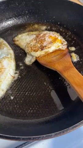 The video captures a close-up view of two fried eggs cooking in a black frying pan. The pan has a distinct hexagonal textured surface. Oil or cooking fat is visible, bubbling gently around the eggs.

At the beginning of the clip, one egg is being flipped by a light brown wooden spatula. The spatula adeptly lifts the egg, which has a well-cooked, golden-brown underside, and smoothly turns it over in the pan. The egg lands back into the hot oil, now showing its other side, which also appears golden brown and slightly crispy. Another fried egg is partially visible on the left side of the frame, already cooked and resting in the pan.

A clear sizzling sound accompanies the cooking process throughout the video. The focus remains tightly on the pan and the cooking eggs, with the wooden spatula being the only other prominent object. The scene suggests a domestic kitchen environment, likely during a meal preparation, focusing solely on the cooking activity.