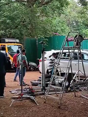 The video shows a film crew preparing for a shoot in an outdoor setting with many trees. Several people are visible, some carrying equipment and others standing around observing. There is a truck, a few cars, and a motorcycle in the background. Film equipment such as cameras, lights, ladders, and various stands are scattered around the set. The ground is covered in dirt and fallen leaves.