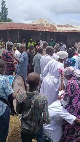 A group of people, many dressed in white, are gathered outdoors. Some are interacting with each other, while others appear to be observing. A large drum is visible in the foreground, with a young person standing behind it. In the background, there is a building with a corrugated metal roof. The overall scene suggests some kind of communal event or gathering.