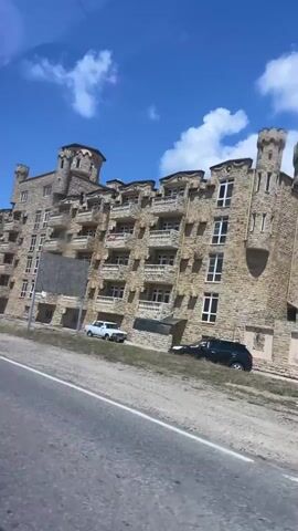 The video shows a roadside view of a building with castle-like architecture. The building has stone facades, turrets, and multiple windows. In front of the building, there are parked cars, including a white SUV and a darker sedan. A sign with text is visible near the building. The scene is set against a bright blue sky with fluffy white clouds. The video appears to be filmed from a moving vehicle, as evidenced by the blurred foreground of a road. There are no people clearly visible in the video.