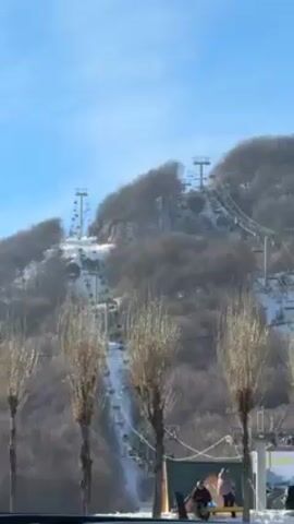 The video shows a ski resort on a sunny day. A ski lift ascends a snow-covered hill, dotted with bare trees. At the base of the hill, near some trees, are a few people sitting on a bench.