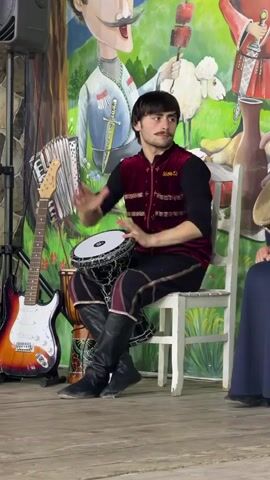A young man with dark hair and a mustache, wearing a dark long-sleeved shirt, a red vest with gold trim, patterned pants, and black boots, sits on a white chair and plays a bongo drum with his hands. He is positioned in front of a colorful mural depicting people, a sheep, and food. To his left, an accordion and an electric guitar are visible. To his right, another person in a dark blue dress is partially visible, holding a drum. The man is looking to his right.