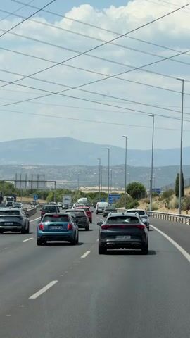 The video shows a view from a vehicle driving on a highway. Several cars are visible in front, moving in the same direction. The road has multiple lanes and is paved with asphalt. Overhead power lines stretch across the sky, supported by tall poles. In the background, distant mountains are visible under a partly cloudy sky. The environment appears to be rural or semi-rural, with vegetation and signs along the side of the road. The lighting suggests it is daytime.
