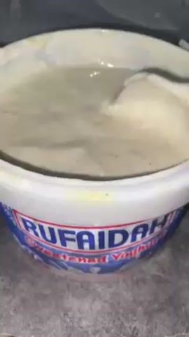 A close-up shot displays a white plastic tub labeled "RUFAIDA" containing a light beige, creamy substance, likely yogurt or a similar dairy product. A white spoon is used to stir and lift the substance, revealing its texture and consistency as it drips back into the tub. The background is blurry, showing a grey surface and a partially visible plastic bag.