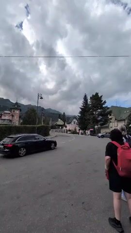 The video shows a cloudy day in a town. A black car is driving on a road. There are a few people standing on the side of the road. The buildings in the background appear to be houses or hotels. There are mountains in the distance. The weather appears to be overcast and potentially rainy.