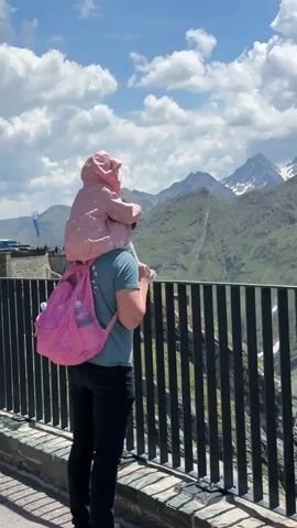 A man wearing sunglasses and a teal t-shirt carries a young child on his shoulders. The child, wearing a pink hooded jacket and white pants with a floral pattern, sits on his shoulders and rests her hands on his head. The man has a pink backpack on his back. They are standing on a walkway with a black metal railing. Behind them, there are mountains, buildings, and a cloudy sky. The man turns his head to look at the camera and smiles. The child looks forward and to the side.