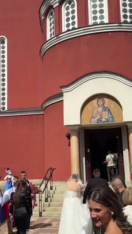 A wedding party approaches a red church with white-trimmed arched windows. A bride in a white dress and veil walks up the stairs toward the entrance. Several guests, dressed in formal attire, follow behind her. A man in a black shirt holds a camera and stands at the entrance, possibly filming the ceremony. The sun shines brightly on the scene.