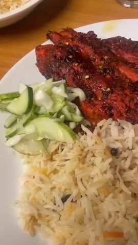 The video shows a close-up shot of a plate of food. The plate contains tandoori chicken, rice, and a cucumber salad. The chicken is a deep red color and appears to be grilled. The rice is long-grained and appears to be seasoned with spices. The cucumber salad is made of thinly sliced cucumbers and possibly some other chopped vegetables. The lighting is warm and appears to be from an overhead source. The background is slightly blurred but suggests a restaurant or home dining setting. There are no people visible in the video. The video is short, with the camera panning slightly across the plate of food.