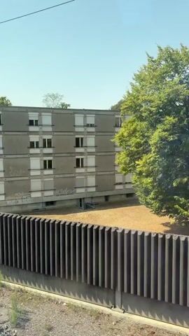 The video shows a view from a moving vehicle, likely a train, as it travels over a bridge. Initially, a concrete apartment building with white-trimmed windows and some graffiti is visible, alongside a tree and a tall, ribbed fence. The perspective then shifts to focus on the underside of the bridge's steel structure, characterized by numerous rivets. Through the gaps in the bridge's framework, a shallow river with a gravelly bed and trees lining its banks can be seen. The sky is clear and blue, indicating daylight.
