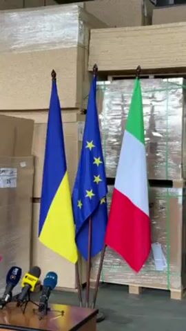 The video shows three flags standing side-by-side in front of stacks of wrapped, rectangular items, likely packages of wood or other construction materials. The flags, from left to right, are the Ukrainian flag (blue over yellow), the European Union flag (blue with a circle of yellow stars), and the Italian flag (green, white, and red). In front of the flags, on a dark brown table, are several microphones from various media outlets, indicated by their logos, including one with a blue logo that says "liv". The scene seems to be set up for a press conference or similar event.