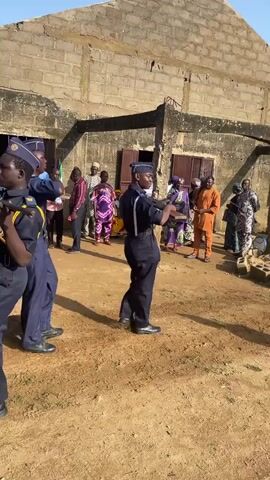 The video shows an outdoor scene in front of a simple brick building. The ground is a sandy, dirt surface. There are several people in the background, some standing and watching. In the foreground, three individuals are dressed in dark blue uniforms with matching caps, appearing to be performing a synchronized march or drill. The two individuals in the center and on the right are facing away from the camera, while the individual on the left is facing towards the building and partially towards the camera. They are all moving their legs in a marching motion. The building has a rough, unfinished appearance with visible brickwork and appears to have doors and windows. Sunlight is casting shadows, indicating it is daytime.