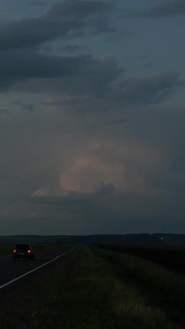 The video depicts a dark, moody scene of a car driving on a road at dusk. The sky is filled with dramatic, heavy clouds, suggesting impending rain or a storm. The lighting is dim, with the car's taillights being one of the few bright elements in the frame. The landscape appears rural, with fields stretching out on either side of the road, bordered by a distant treeline. The overall atmosphere is somber and quiet, evoking a sense of solitude or journey. The time of day is clearly evening, as indicated by the low light and the appearance of the sky. There are no people visible, only the solitary car and the vast, atmospheric landscape. No text is visible in the video.
