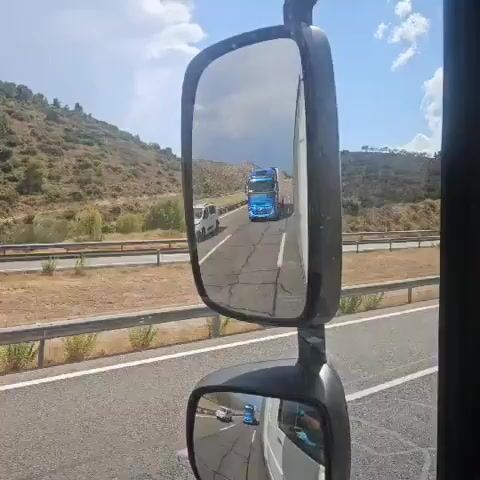 The video shows a close-up view of a truck's side mirror. The mirror reflects a bright blue semi-truck driving on a highway. The highway appears to be in a dry, hilly, or mountainous region. The sky is visible in the reflection, with some clouds. There is also a smaller white vehicle visible in the lower mirror. The video appears to be filmed from another vehicle driving on the same highway.