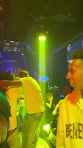The video shows a crowded nightclub with people dancing and sitting. A man in a white shirt and light-colored jeans dances in the foreground. Behind him, some people are seated at tables. A screen displays a video, showing a person in a similar outfit as the dancer. The camera focuses on a young man wearing a yellow hoodie with the word "BENEVOLENT" written on it. He initially smiles and then looks down. There are bright green and red lights flashing in the club, contributing to the lively atmosphere. The venue seems to be a restaurant and bar as well, denoted by the sign in the corner. A man can be seen playing percussion instruments in the background.