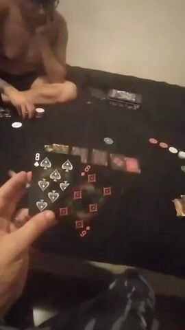 Two people are sitting on a dark surface, possibly a bed, playing cards. In the foreground, a hand holds an 8 of spades and a 6 of diamonds. There are several other cards and poker chips scattered on the surface. One person in the background appears to be looking at the cards. The environment is dimly lit.