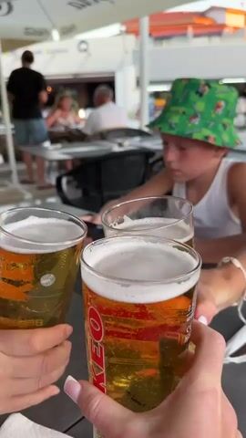 The video shows a group of people at an outdoor cafe. In the foreground, three glasses of beer are being raised in a toast. The glasses are clear and filled with golden-colored beer with a frothy head. One glass has the word "KEO" printed on it, along with "1951" and "FINEST QUALITY BEER." The people holding the glasses are only partially visible, with hands and manicured fingernails in focus.

In the background, a young boy wearing a green bucket hat with cartoon characters is seated at a table. He is looking down, and his face is slightly blurred. Further in the background, other patrons are visible, sitting at tables and appearing to dine or socialize. The setting is an outdoor seating area with umbrellas providing shade. Beyond the cafe, a street scene is visible with buildings and shops, suggesting a commercial or tourist area.

The lighting suggests it is either late afternoon or early evening, with a warm, soft light illuminating the scene. The overall mood appears relaxed and convivial, centered around enjoying drinks in a social setting. The location context of Peyia, Cyprus, combined with the visible brands and general atmosphere, points to a holiday or leisure setting.