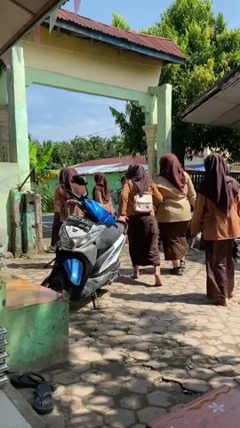 A group of five female students wearing brown uniforms and hijabs are standing near an arched entryway. One student, in the foreground, is walking away from the camera and appears to be holding the hand of another student. A blue and silver scooter is parked to the left of the students. The ground is paved with interlocking bricks, and a building with a red roof is visible behind the entryway. Trees and a clear sky are present in the background. The students are interacting with each other and moving in the same direction.