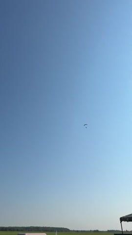 The video begins with a clear blue sky. A paraglider can be seen in the distance. A person's hand, wearing a blue and white long-sleeved shirt, enters the frame from the right, pointing towards the sky. The paraglider continues to fly. In the background, there is a flat, green field with some structures that appear to be part of an airfield, including a large white cylindrical object, a tent-like structure, and a bench. Another paraglider appears closer to the foreground. The camera pans slightly to the right. The paraglider in the distance is still visible, and another paraglider is seen closer. The video continues to show the paragliders against the blue sky and the airfield in the background.