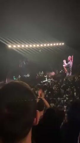 The video shows a concert scene from an elevated perspective. A singer stands center stage, illuminated by spotlights, wearing a dark shirt and light jeans. Behind him are three men dressed in black and a guitarist. The stage is decorated with minimalistic arch structures. Large screens flank the stage, projecting close-up images of the singer. A dense crowd of people are visible in the foreground, many with their arms raised. The overall lighting in the venue is dim, emphasizing the bright stage.