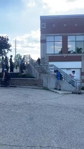 A group of individuals are gathered in a paved outdoor area in front of a brick building with large windows. Several people are standing near a set of outdoor stairs, some of whom appear to be holding cameras or phones. One individual on a scooter is riding down the stairs and appears to have fallen. Another individual is on the ground in the foreground, appearing to recover from a fall. The sky is blue with some clouds, and there is greenery visible to the left of the building.