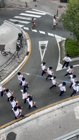 A high-angle, full shot captures a street parade. A large group of young people, mostly male, clad in white shirts and dark pants, march in formation down a street. They are playing drums, some of which are small and held by hand, while others appear larger and possibly strapped to their bodies. The street is paved and lined with buildings. A pedestrian crossing with white stripes is visible in the background. Several adults and a bicycle are also present on the sidewalks and street. The parade appears to be moving from the foreground towards the background. Blue and white banners are strung across the street above the marchers. The lighting suggests it is daytime, possibly overcast. The overall scene depicts a festive or ceremonial event.