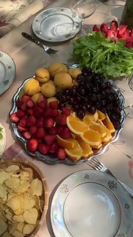 The video shows a close-up overhead shot of a picnic spread. A silver platter is filled with an assortment of fresh fruits, including strawberries, apricots, cherries, and orange slices. A person's hand is visible in the lower left of the frame, reaching for a strawberry. To the left of the fruit platter, a basket of potato chips is visible. To the right of the fruit platter, there are plates, glasses, and a small arrangement of greenery and tomatoes. The setting appears to be outdoors, with dappled sunlight suggesting a natural environment.