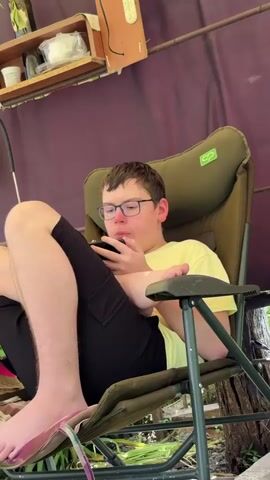 A young male with short brown hair and glasses sits in a green camping chair, wearing a light yellow t-shirt and black shorts. He holds a black smartphone with both hands, looking at the screen. His left foot rests on the chair's metal frame, while his right foot is tucked under his left knee. Behind him, a purple fabric screen is visible, with a wooden shelf holding various items above it. He occasionally moves his lips.