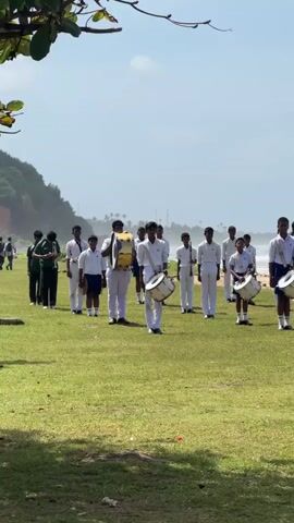 A group of young males in uniform, some carrying drums and others holding what appear to be wind instruments, stand in formation on a grassy area. They are positioned in front of a beach with waves visible in the background under a bright sky. The individuals are mostly facing forward, and some appear to be playing their instruments. The scene suggests an outdoor performance or practice by a school band.