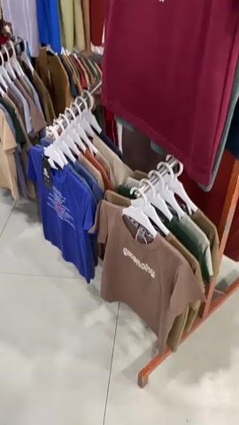 The video pans across a retail store displaying racks of clothing. The items are predominantly t-shirts and some hoodies, with a variety of colors and designs. Several t-shirts feature prominent brand logos such as Fendi, Givenchy, and Balenciaga. The store appears to be well-stocked, with clothes hung neatly on hangers. The floor is tiled, and the lighting suggests an indoor environment. There are no people visible in the video. The camera movement is smooth, giving a comprehensive view of the merchandise. The overall impression is of a clothing boutique or a section of a larger department store.