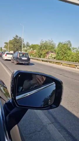 The video shows a view from inside a moving car, focusing on the side-view mirror and the road ahead. In the side-view mirror, a person's reflection is visible, holding a phone and appearing to film. The road ahead is lined with trees and some vegetation, with a clear blue sky above. Several cars are seen on the road, either driving in the same direction or in the opposite lane. The cars include a black station wagon, a white hatchback, a dark colored hatchback, and a silver hatchback. Traffic appears to be moderate, with cars spaced relatively close together. The scene suggests a sunny day, possibly in a suburban or rural area with good road infrastructure.