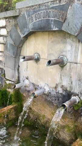 The video shows a water fountain with six metal pipes jutting out of a stone wall. Water flows from the lower three pipes, creating small waterfalls into a mossy basin below. Above the pipes, the words "СВ. БОГОРОДИЦА" are inscribed in a stone arch. The wall is composed of irregular stone blocks and appears weathered and aged. The surrounding environment is green, suggesting an outdoor, possibly natural setting. The water appears clear and clean. The footage is stable, providing a clear view of the structure and water flow.