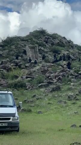 The video shows a rocky hillside in a green, grassy field on a cloudy day. On the hillside, a stone-like structure resembling a doorway is partially visible. Several people dressed in black outfits are seated on the rocks, possibly actors in a scene. Further up the hill, there are what appears to be film crew members, along with lighting equipment, a camera stand, and possibly a boom mic. A white van is parked in the foreground. The scene suggests a movie or film being shot in a rocky, natural setting.