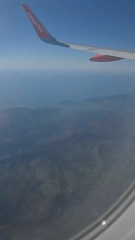 The video is an aerial view from an airplane window looking down at land and sea. An airplane wing with the orange and white EasyJet logo is visible in the upper portion of the frame. Below the wing, the sea stretches to the horizon, meeting a clear blue sky. The land below appears hilly and appears to be somewhat developed with some structures visible, possibly a town or village, near the coast. The terrain is a mix of darker vegetated areas and lighter patches, possibly indicating roads or cleared land. The view is slightly hazy, suggesting distance or atmospheric conditions. The camera is stationary, capturing a static scene.