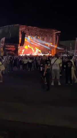 The video shows an outdoor concert at night. A stage is set up with bright lights and a band performing. A large crowd of people is gathered in front of the stage, watching the performance. Some people are standing, while others are sitting on the ground. The atmosphere appears lively and energetic.