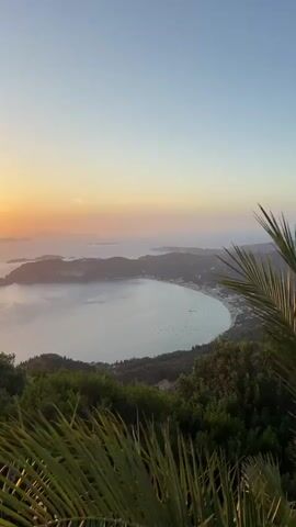 A video taken from an elevated viewpoint shows a wide bay and a coastline at sunset. The sun is low on the horizon, casting a warm orange glow. Several small islands are visible in the distance. The foreground is filled with lush green foliage, including trees and palm fronds. The water in the bay is calm and reflects the colors of the sky. A few boats are anchored in the bay, and some buildings are visible along the coastline. The overall scene depicts a serene and picturesque coastal landscape at dusk.
