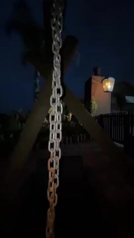 At night, a metal chain hangs in the foreground. Behind it, a wooden structure with crossed beams is visible. A lighted lantern sits atop a building, and a wooden fence runs along the property line. There are some trees and foliage visible as well. The overall lighting is dim, making the scene appear dark. The camera moves slightly.
